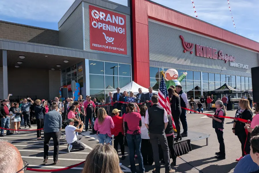 Mayor Tracie Crites at the outdoor grand opening ceremony and ribbon cutting for the new King Soopers Marketplace in Frederick, Colorado, standing at a podium with the American flag and community members gathered around.